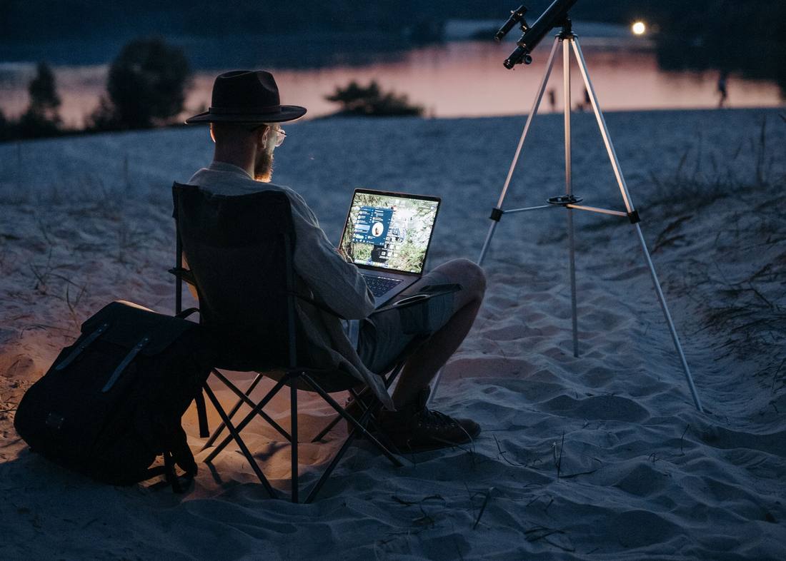 A person working at a laptop beside a telescope on a beach at dusk